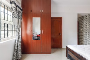 A bedroom with a wooden wardrobe, a bed, and a window with patterned curtains. - The Crimson Cove (Bengaluru)