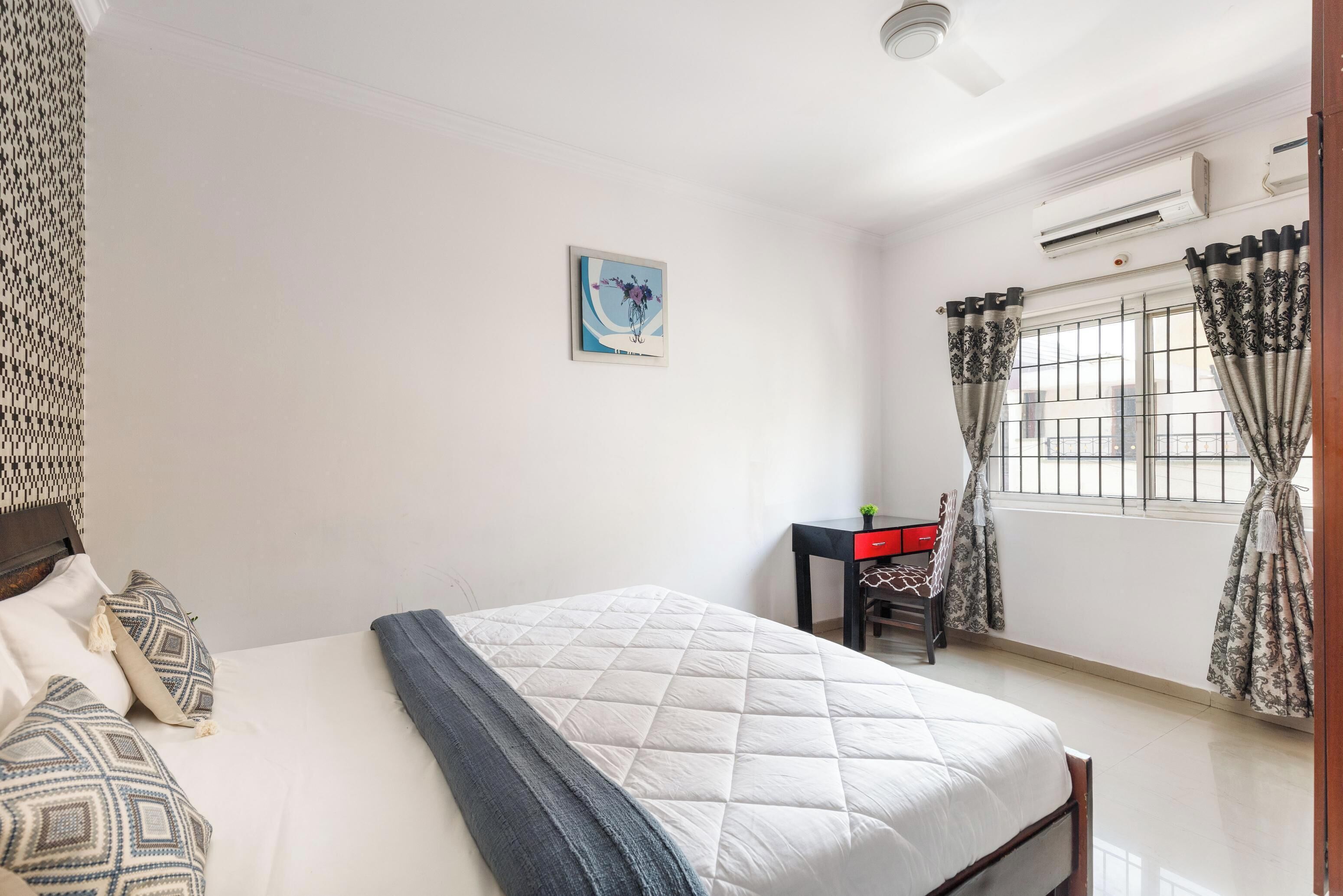 Un dormitorio con cama, escritorio, silla, ventana con cortinas y aire acondicionado.