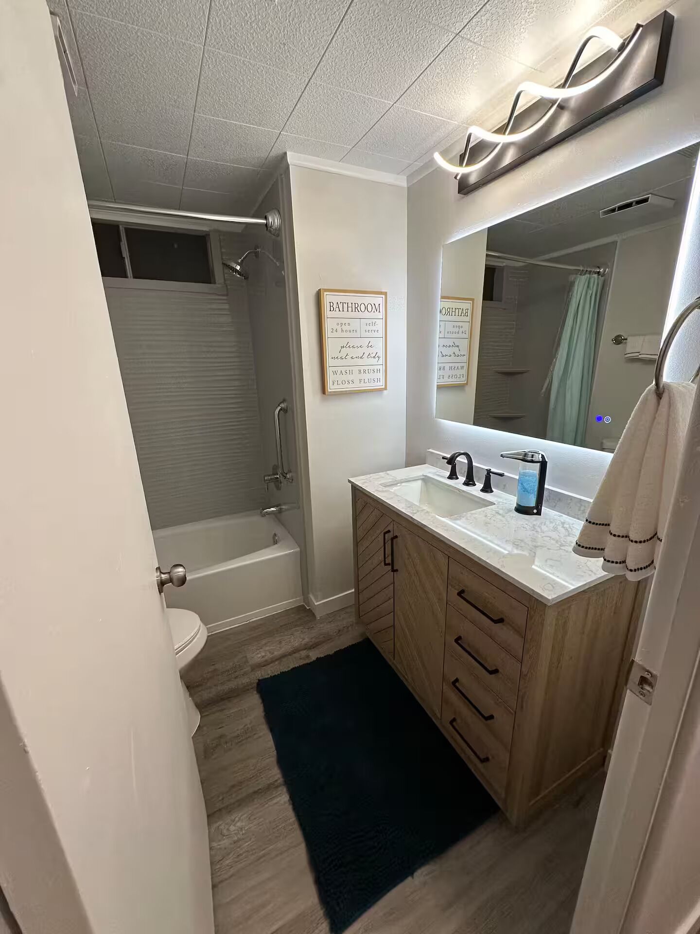 A bathroom with a bathtub, a shower, a sink with a mirror, and a toilet. There are signs on the wall indicating the bathroom is open 24 hours and providing instructions.