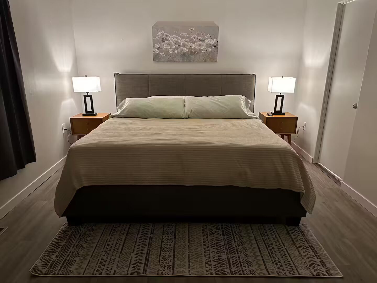 Ein ordentlich bezogenes Bett mit Kopfteil, zwei Nachttische mit Lampen, einem gemusterten Teppich und einem floralen Gemälde an der Wand.