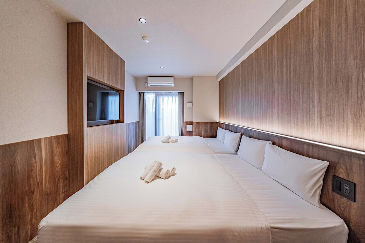 A hotel room with two beds, wooden paneling, a TV, and a window with curtains.