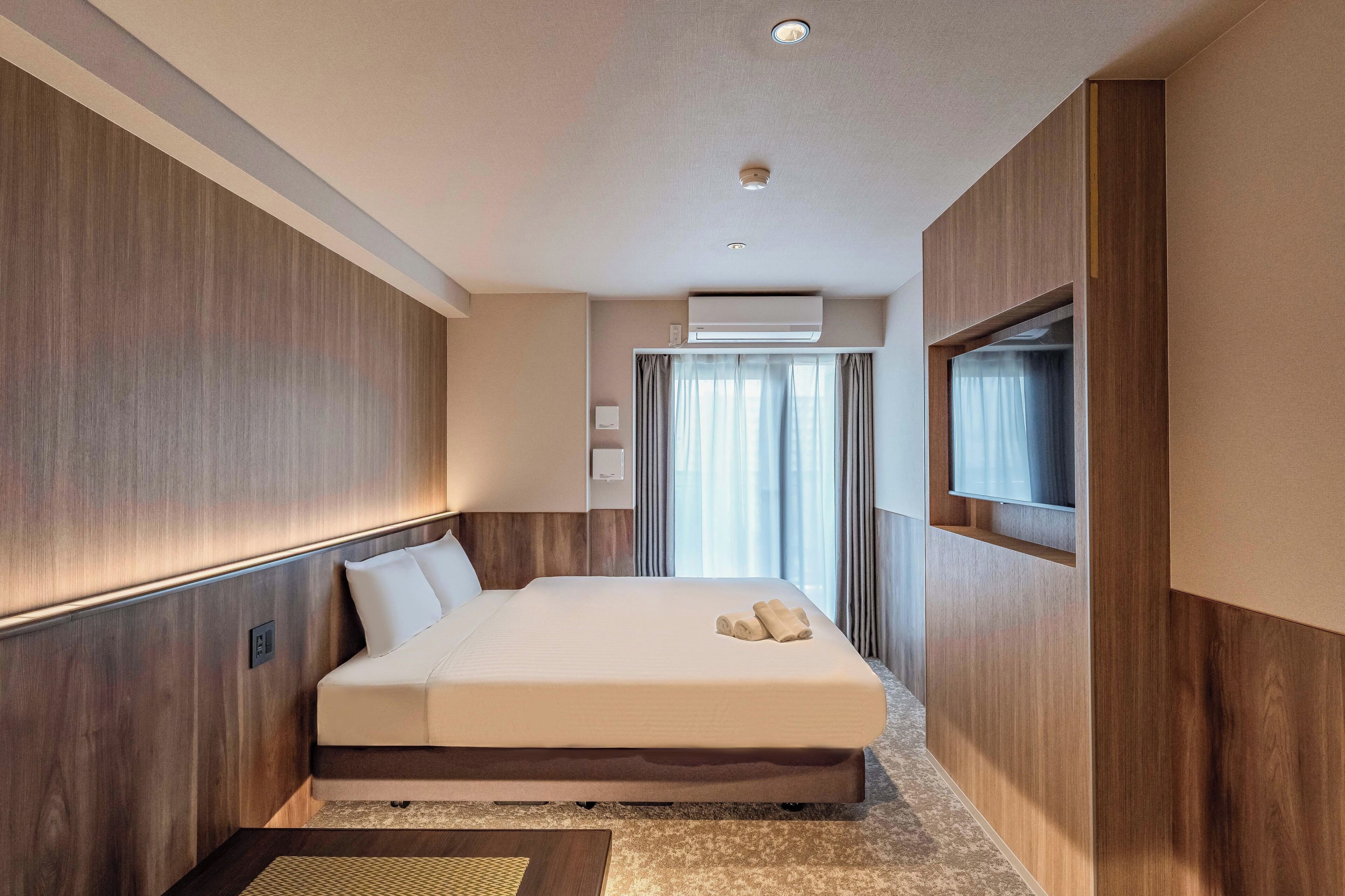 A modern hotel room with a large bed, wooden paneling, a flat-screen TV, and an air conditioning unit.