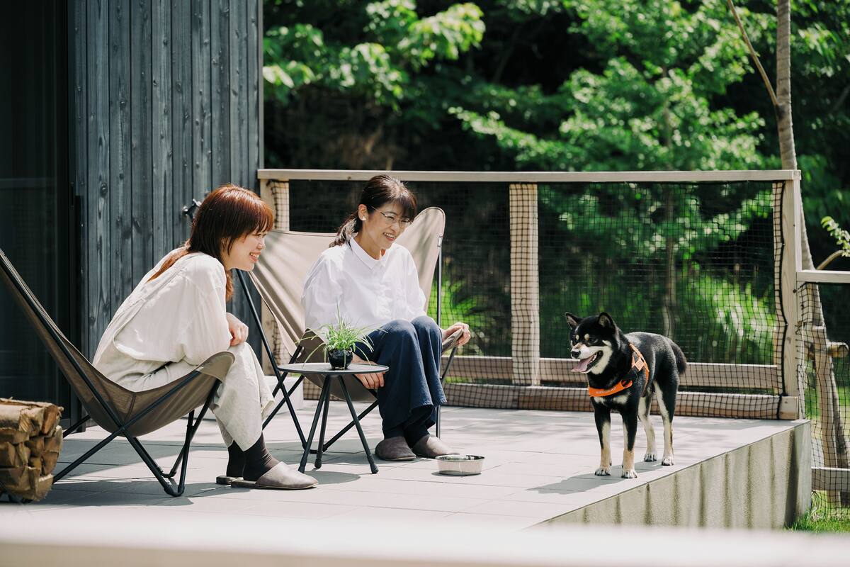 Deux personnes sont assises sur des chaises à l’extérieur, devant une petite table, et une plante en pot est placée entre elles. Un chien se tient sur une corniche au fond.