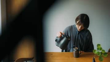 A person watering a plant at a wooden table with a dark mug and a small vase.