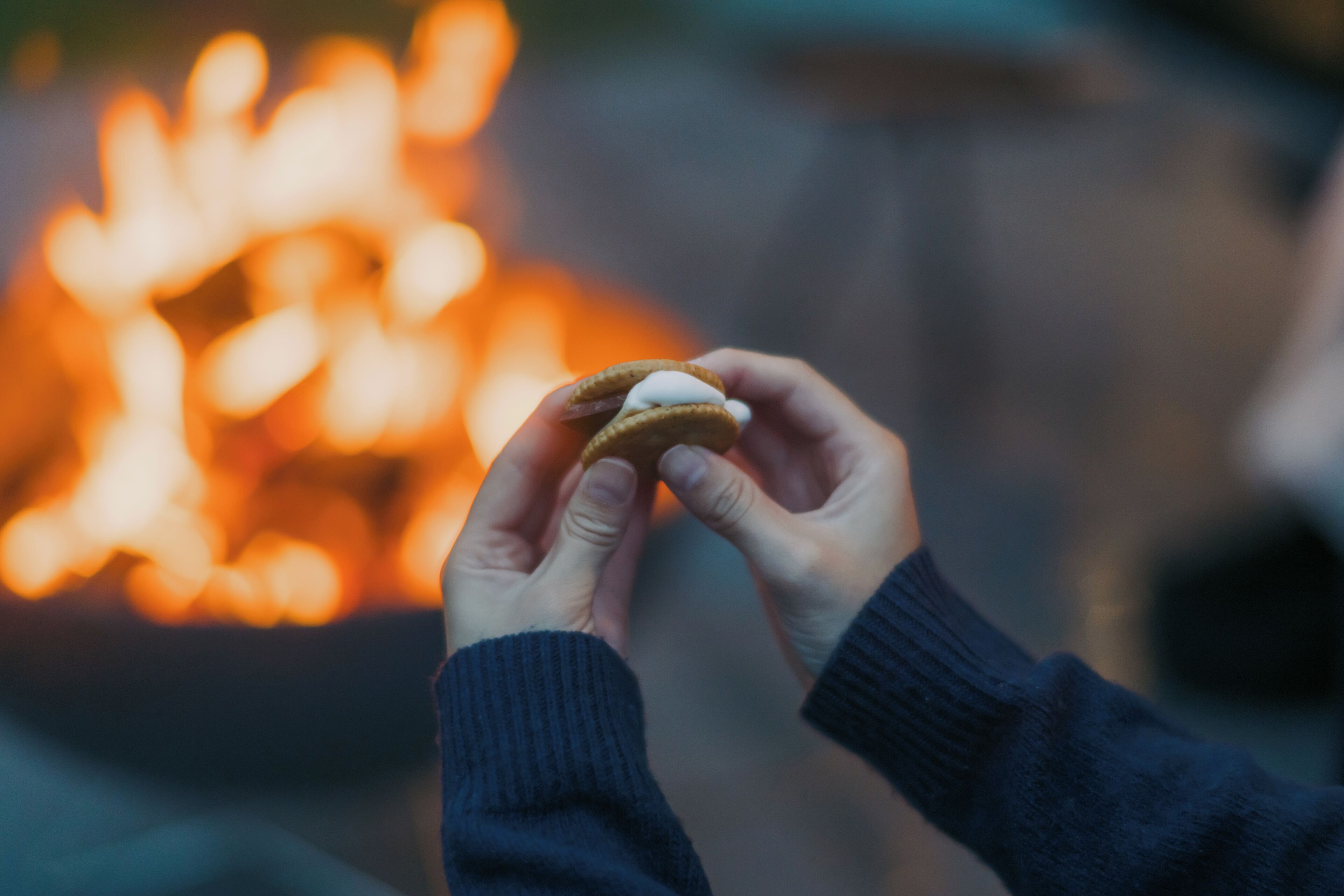 Una persona sostiene una galleta con un malvavisco frente a un fuego desenfocado.