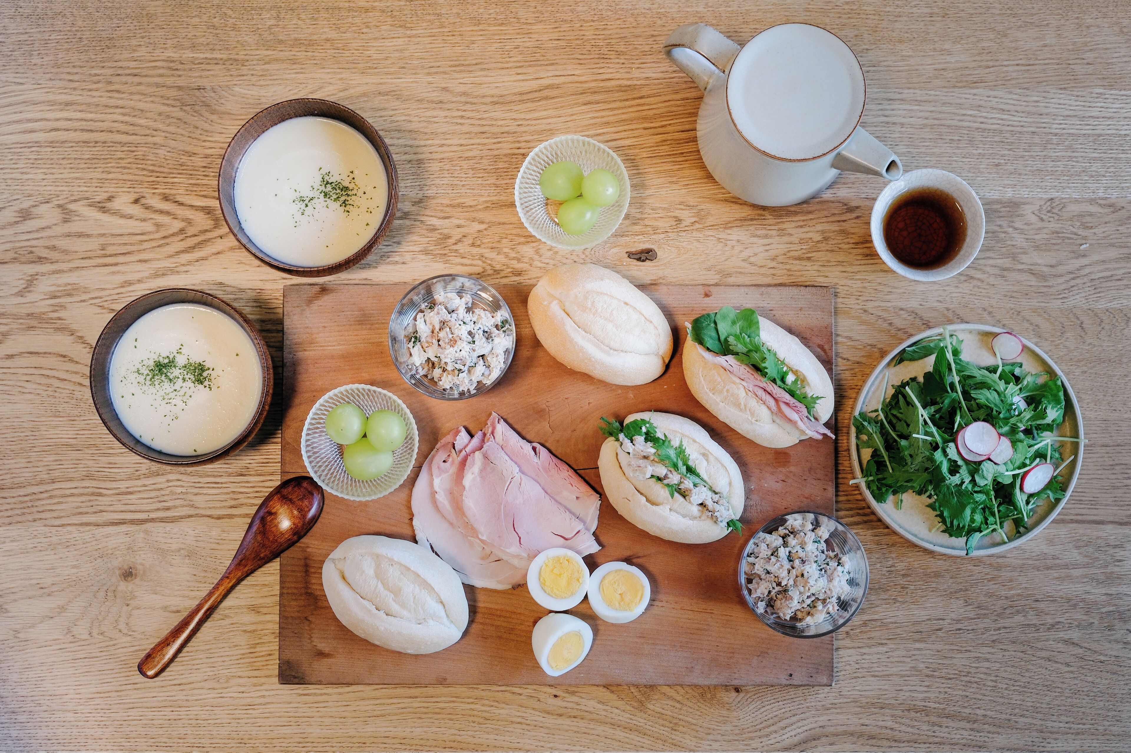 Een houten tafel met diverse etenswaren, waaronder soepkommen, een saladekom, plakjes ham en brood.