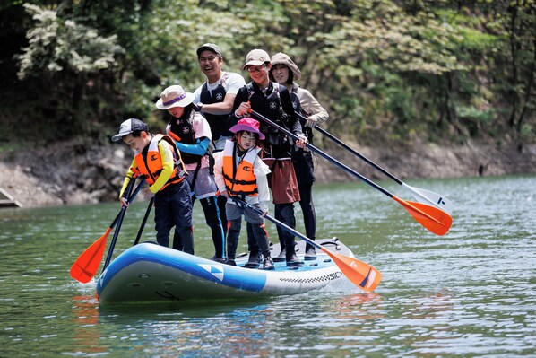 A group of people on a blue and white inflatable raft with orange paddles, standing on a calm body of water surrounded by trees. - 山座熊川 Sanza-Kumagawa (Wakasa)