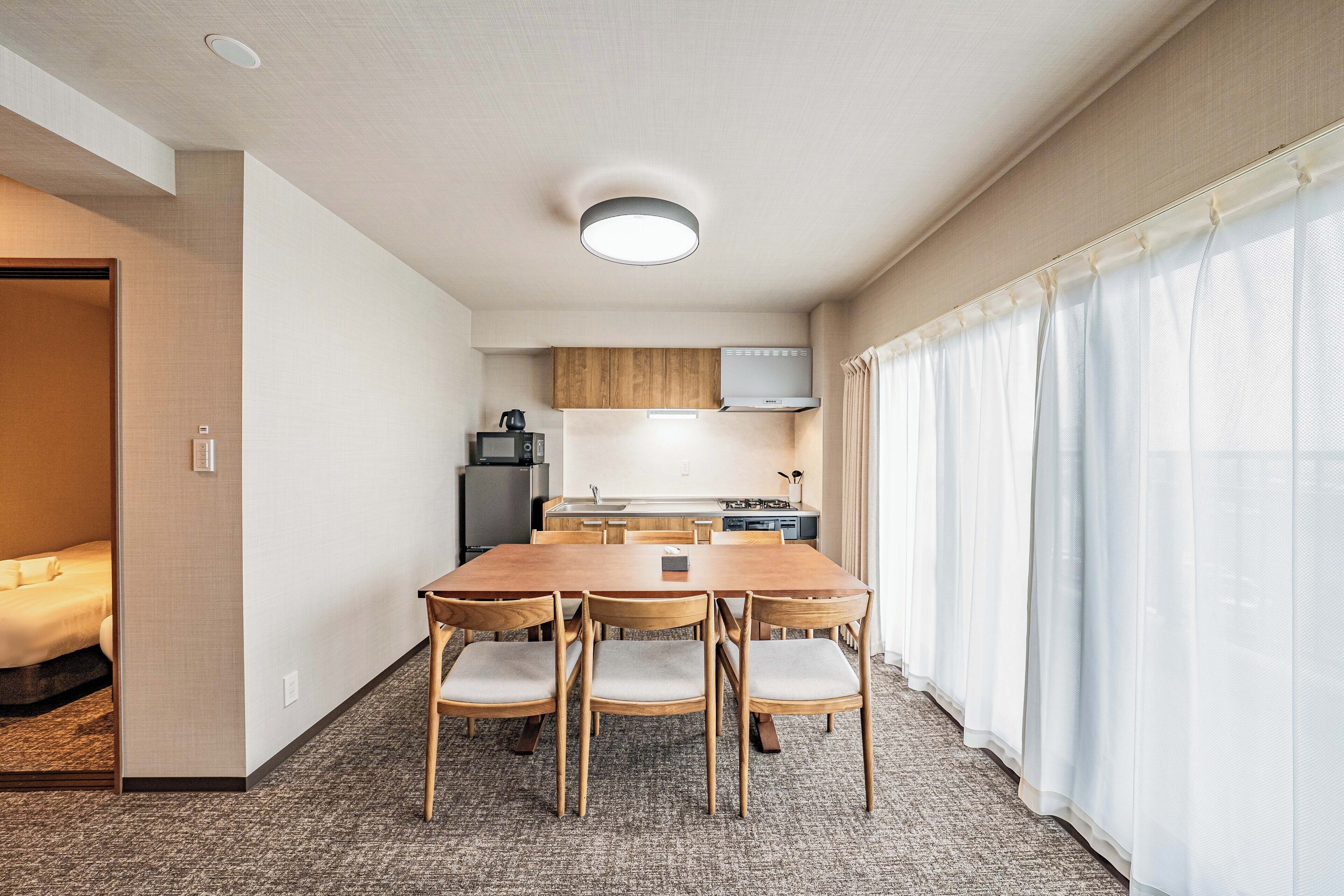 Kompakti hotellihuone, jossa on puinen ruokapöytä ja -tuolit, pieni keittiönurkkaus ja sänky.