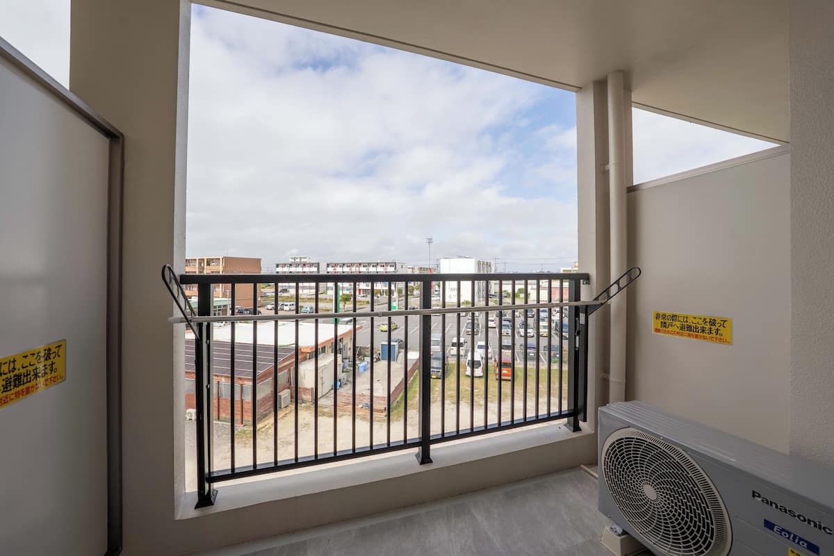 Ein Balkon mit Blick auf einen Parkplatz und Gebäude, einem schwarzen Metallgeländer und einer Panasonic Klimaanlage.