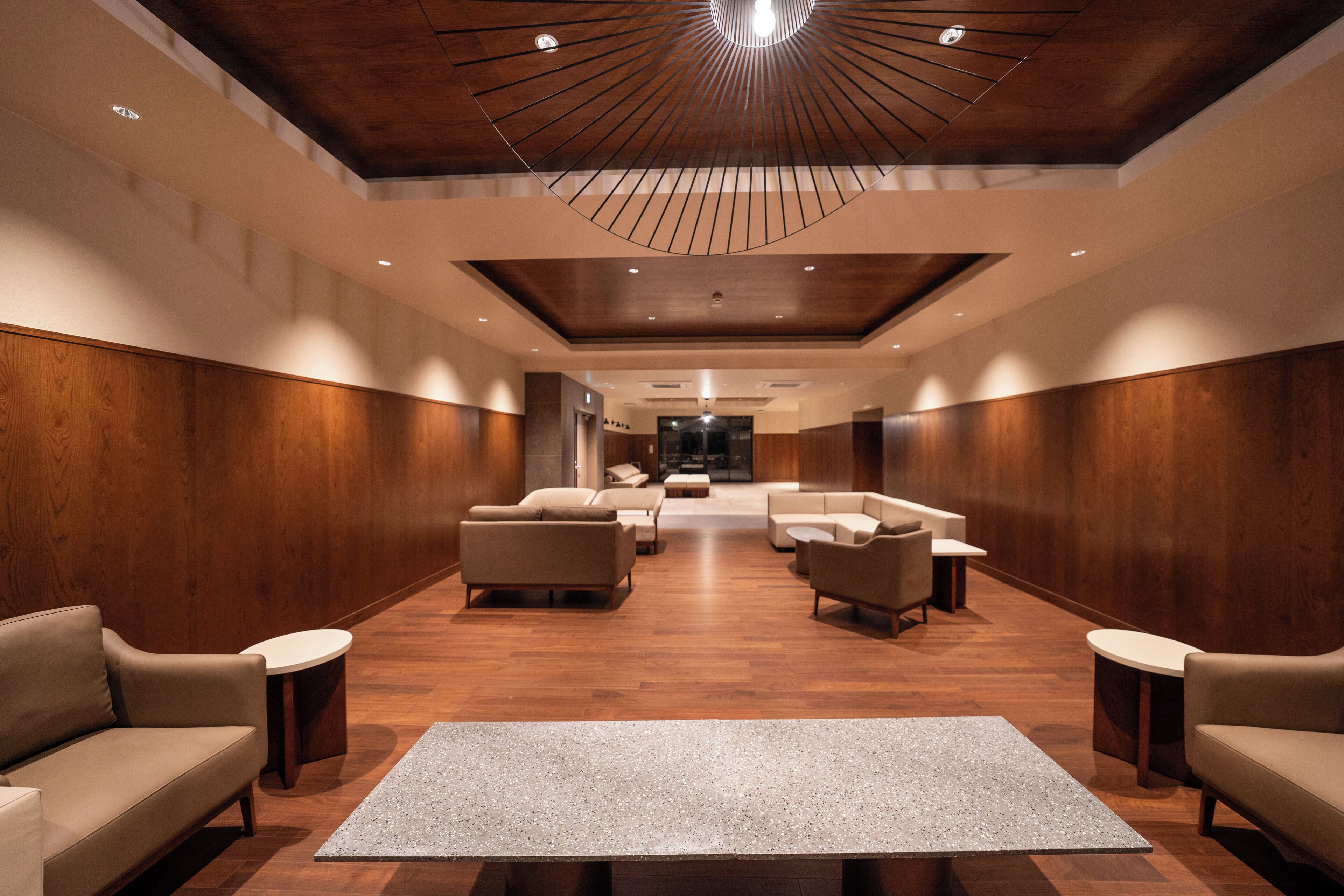 Un área de estar moderna con paneles de madera, una zona central con sofás y una mesa de centro, y un techo con una lámpara radial.