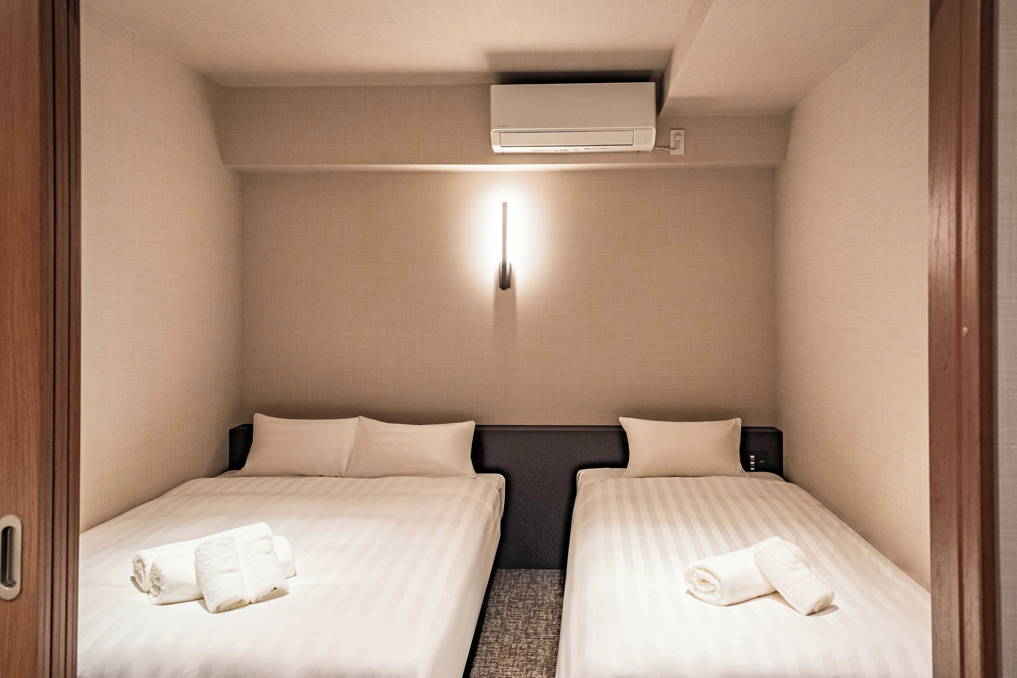 A compact hotel room with two beds, a wall-mounted air conditioner, a bedside table, and a nightstand.