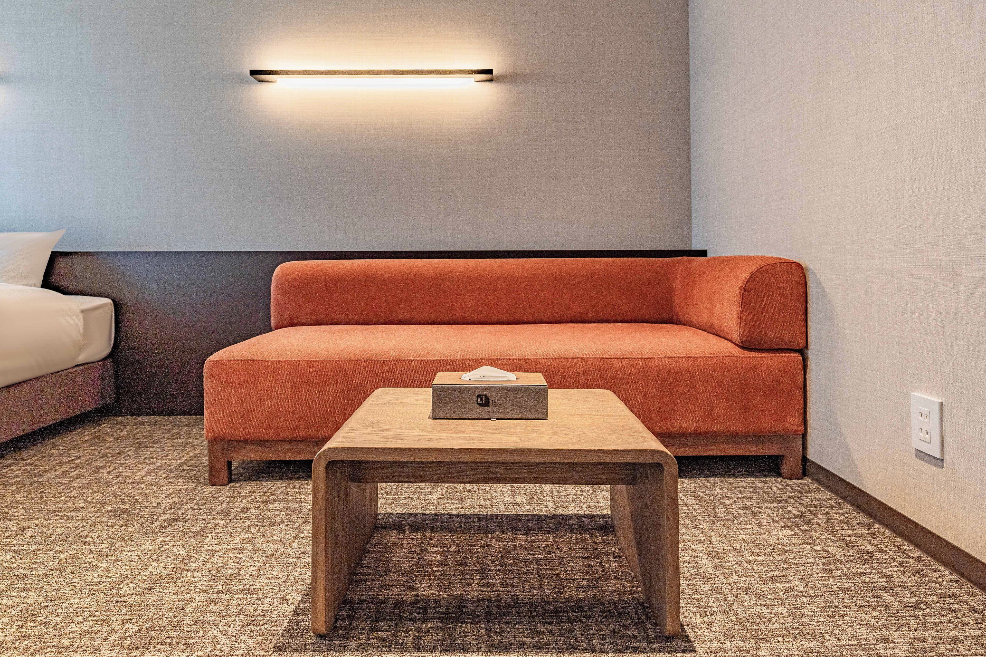 A modern hotel room with a sofa, a small wooden table, and a wall-mounted light fixture.