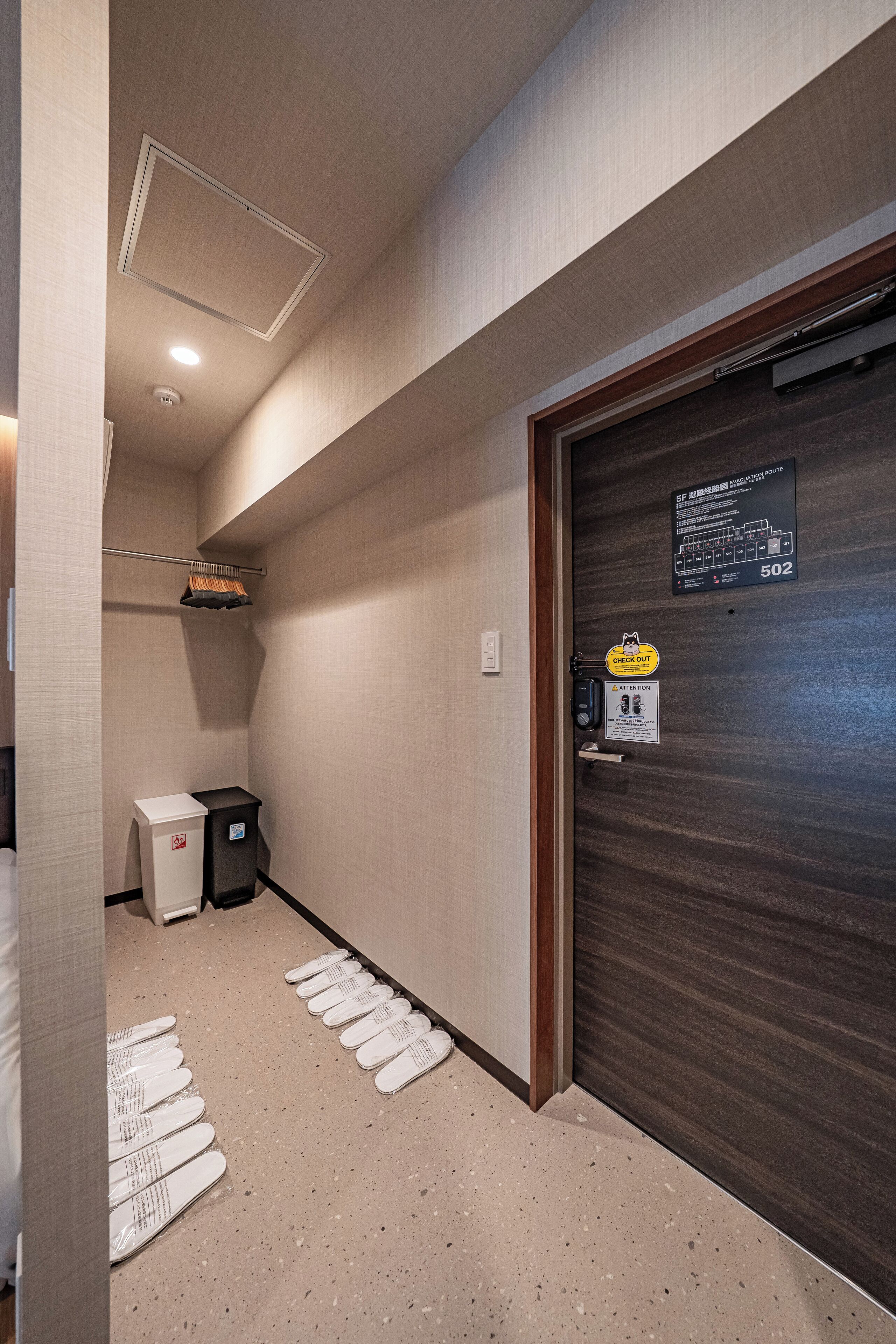 A hotel hallway with a wooden door marked 502, a coat rack, a trash bin, and a row of white slippers on the floor.