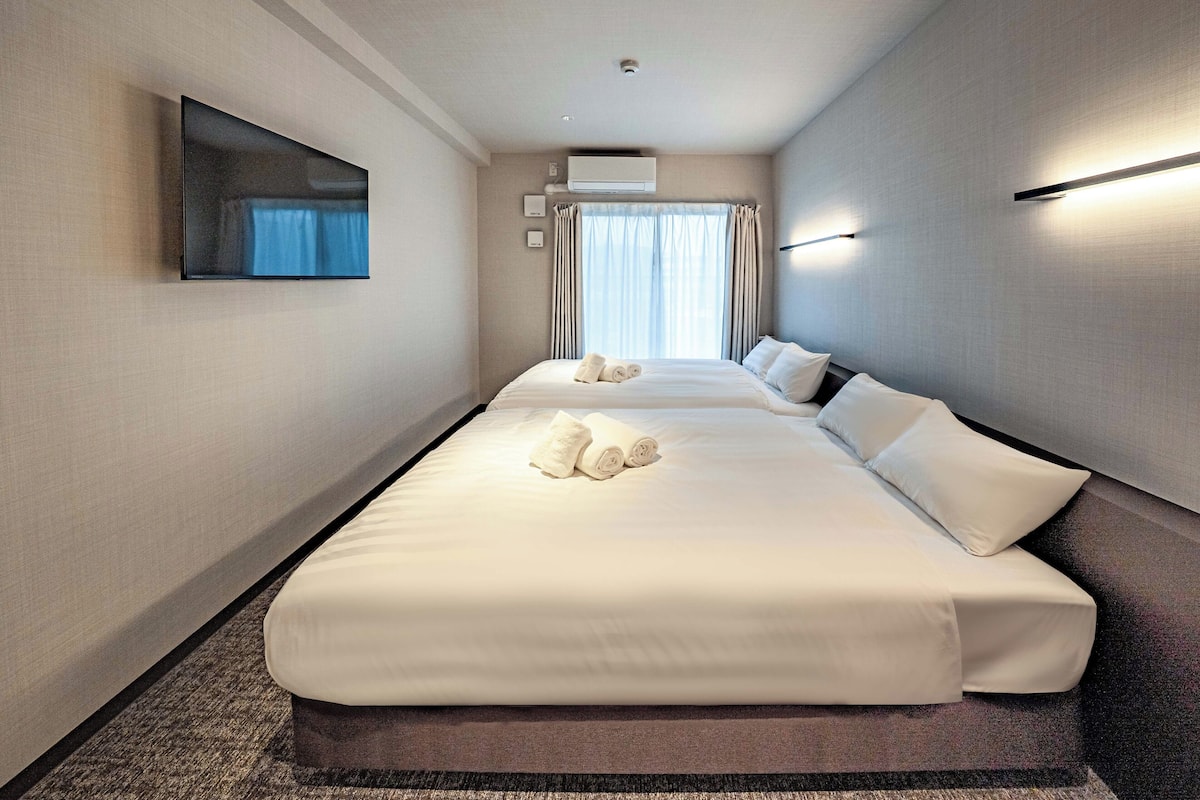 Ein Hotelzimmer mit zwei Betten, einem an der Wand montierten Flachbildfernseher, einer Klimaanlage und einem mit Vorhängen versehenen Fenster.