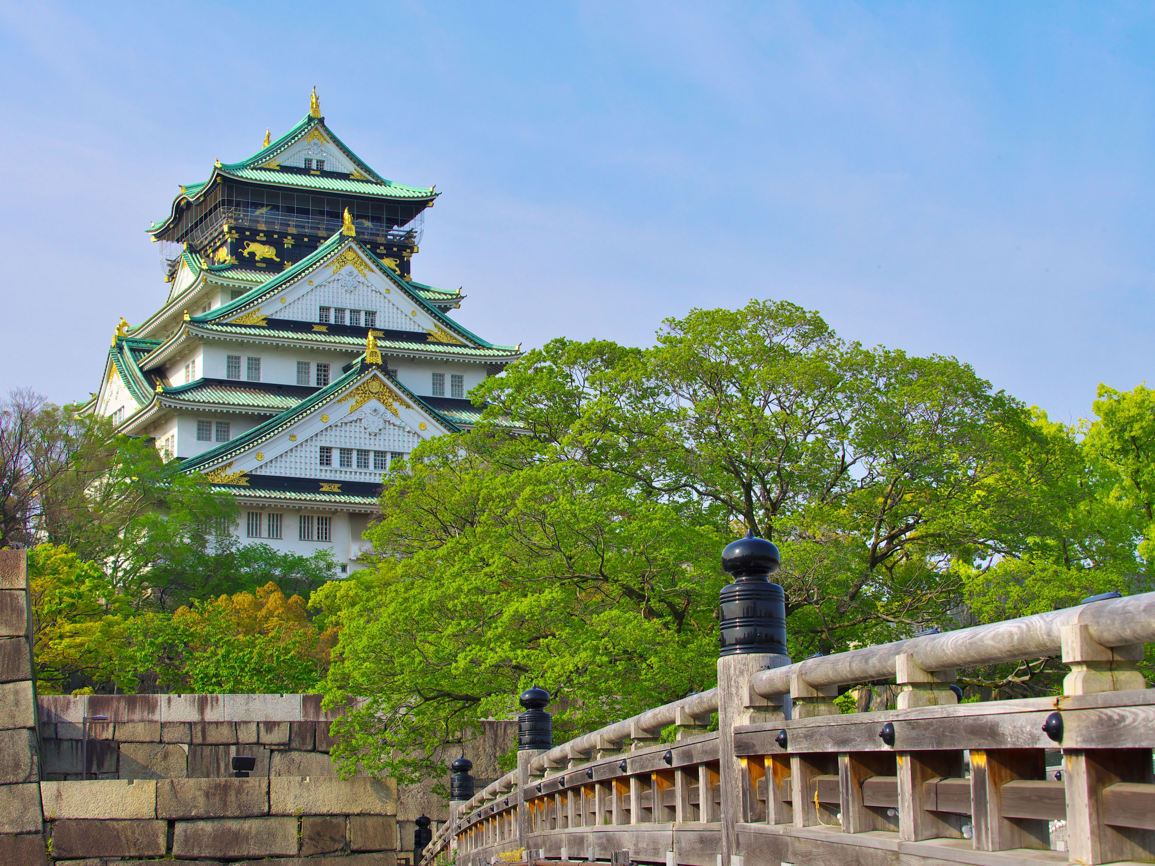 A traditional Japanese castle with multiple tiers and green roofs, surrounded by lush greenery and a stone bridge.