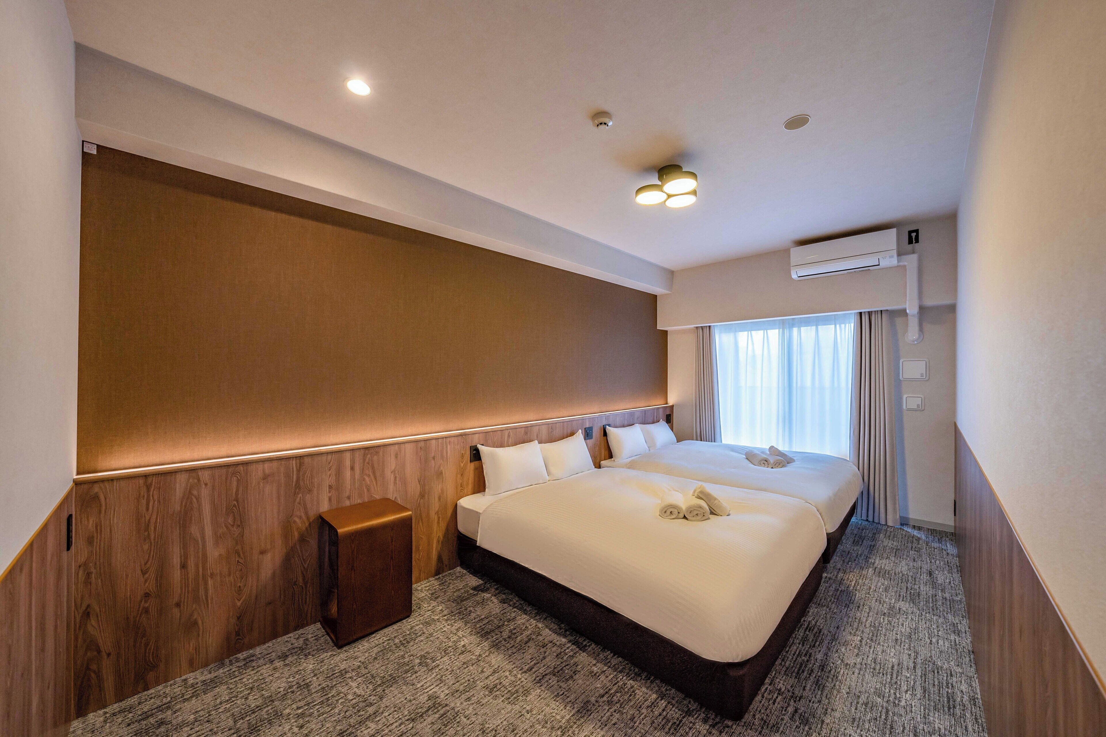A hotel room with a large bed, a wooden headboard, a small wooden side table, and a ceiling-mounted air conditioning unit.