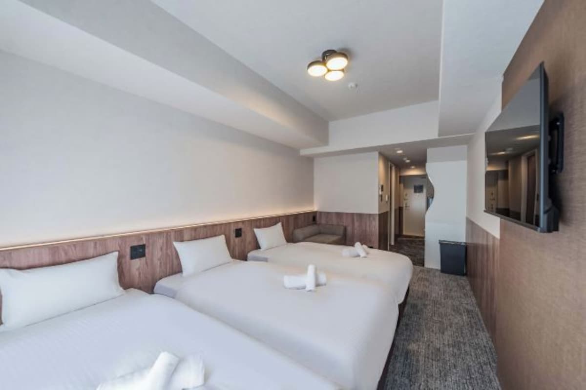 Ein Hotelzimmer mit zwei Betten, einem an der Wand befestigten Fernseher und einem Flur zu weiteren Zimmern.