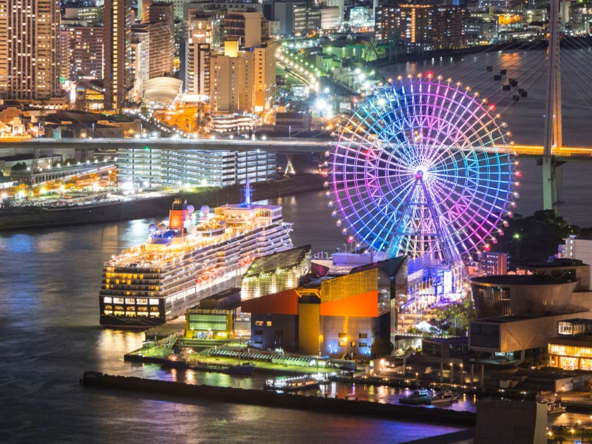 A waterfront cityscape with a brightly lit Ferris wheel and a cruise ship docked at a port.