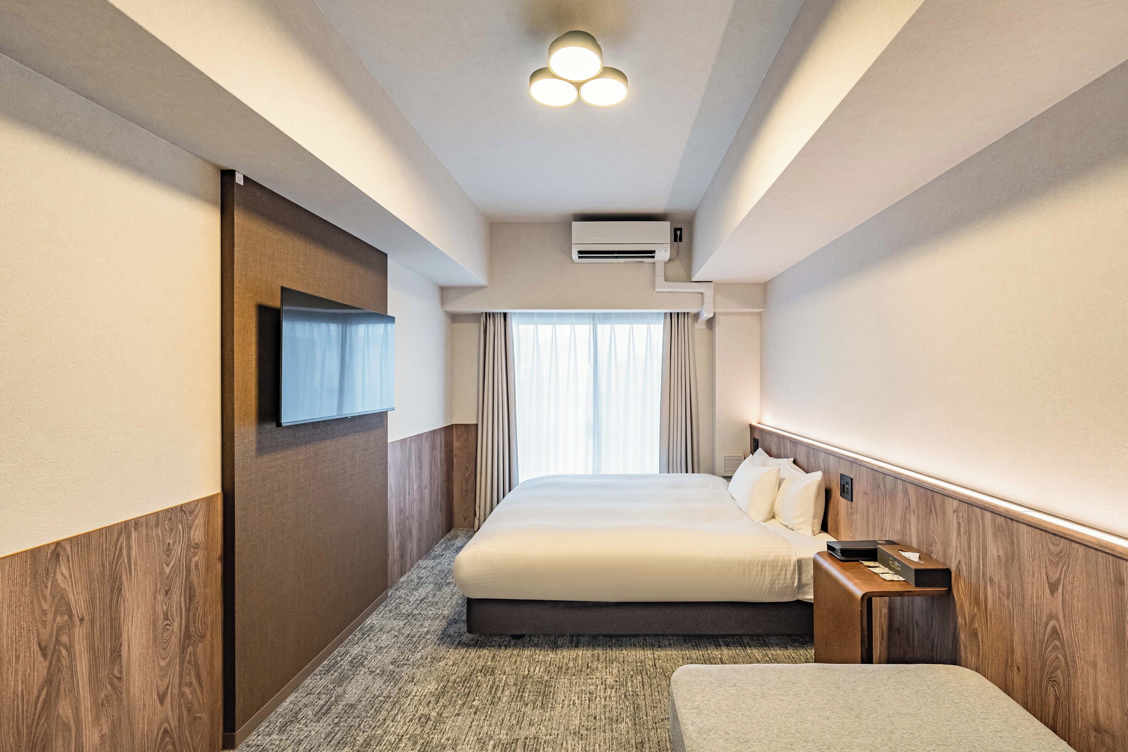 A hotel room with a large bed, a flat-screen TV mounted on the wall, an air conditioning unit, and a window with curtains.