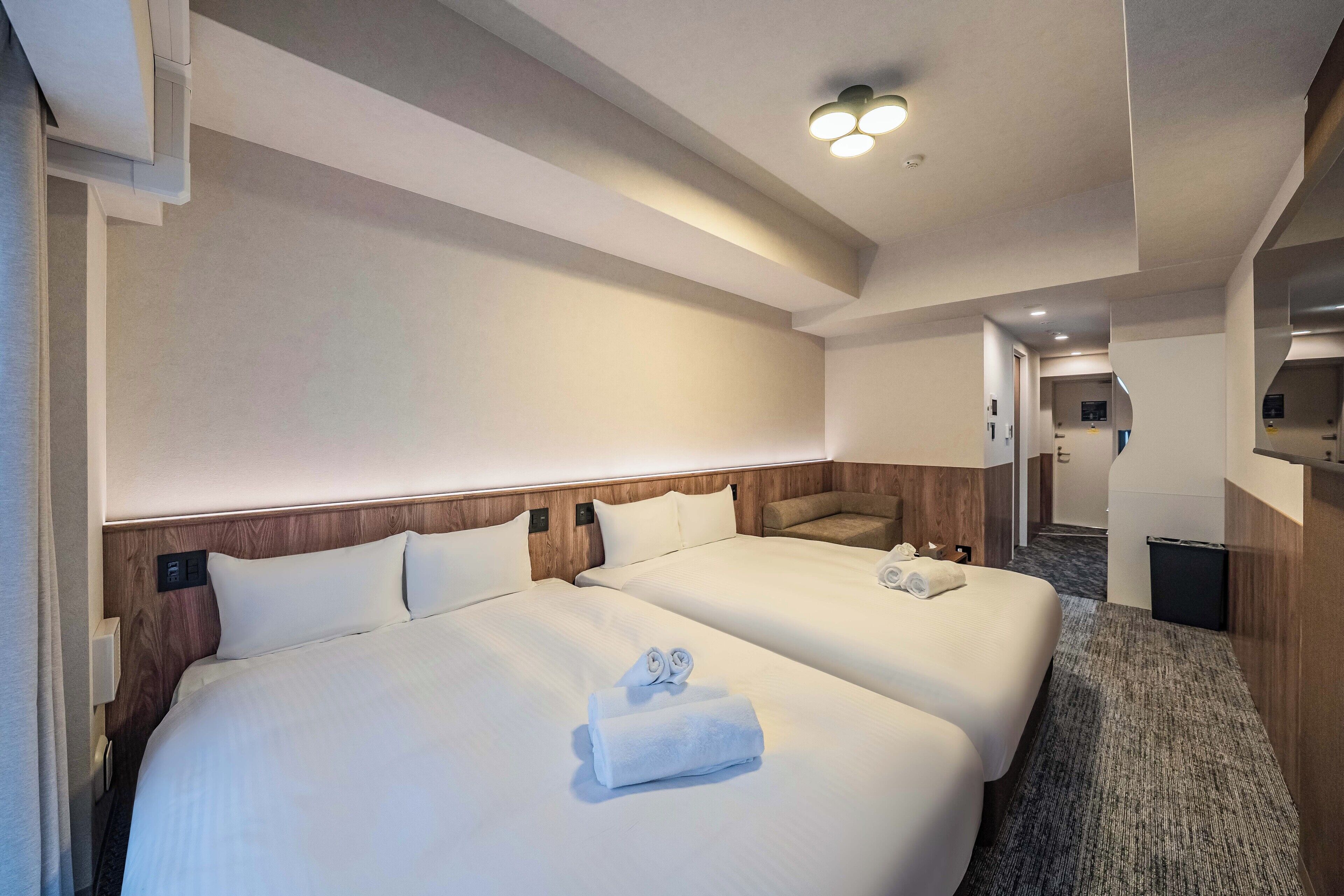 A hotel room with two beds, a wooden headboard, a carpeted floor, and a hallway leading to a bathroom.