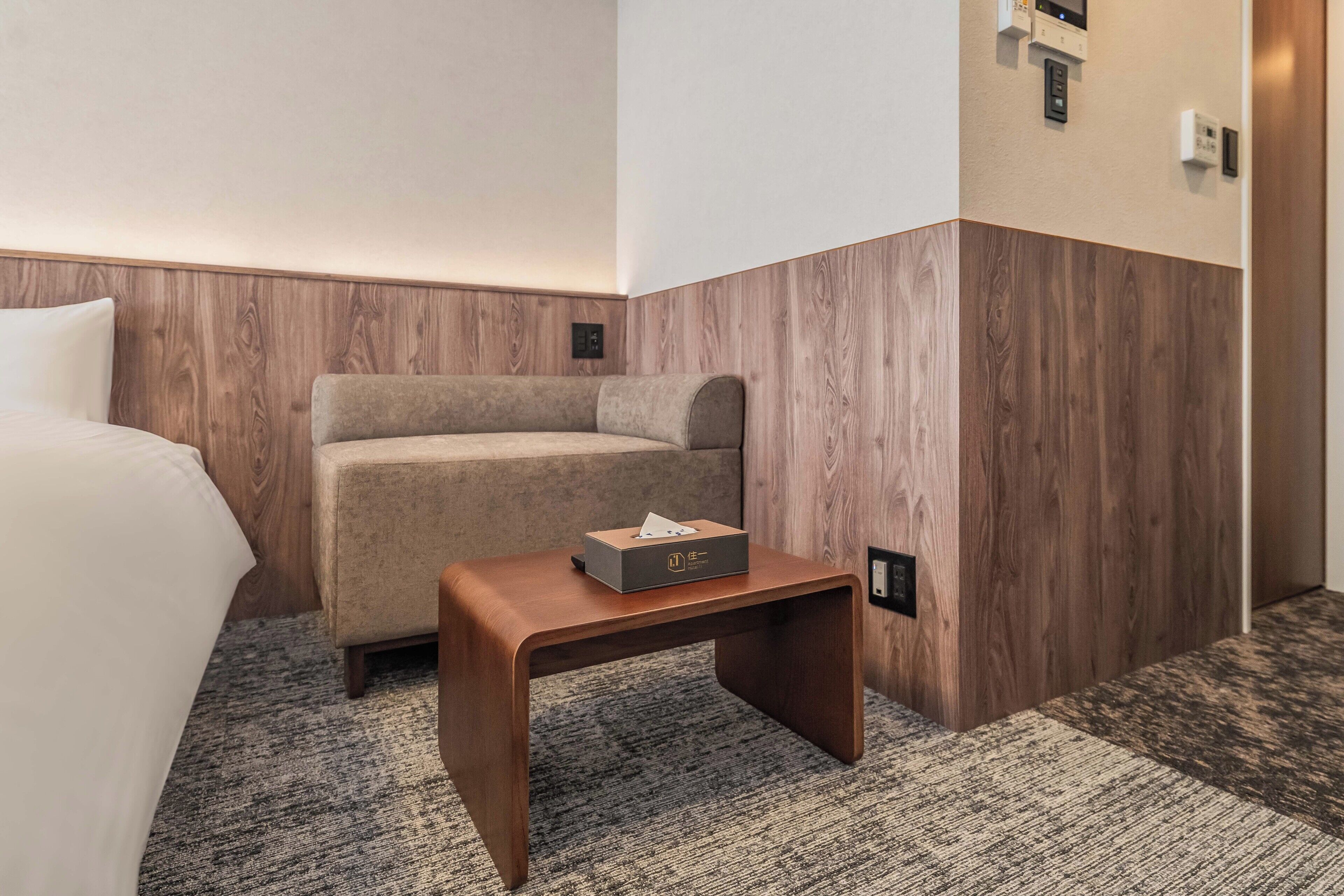 A hotel room with a bed, a sofa, a small wooden table with a tissue box, and a wall-mounted thermostat.