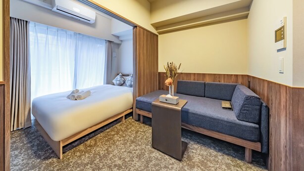 A hotel room with a bed, a sofa, a small table, and an air conditioning unit. - Apartment Hotel 11 Higashi Umeda (Osaka)
