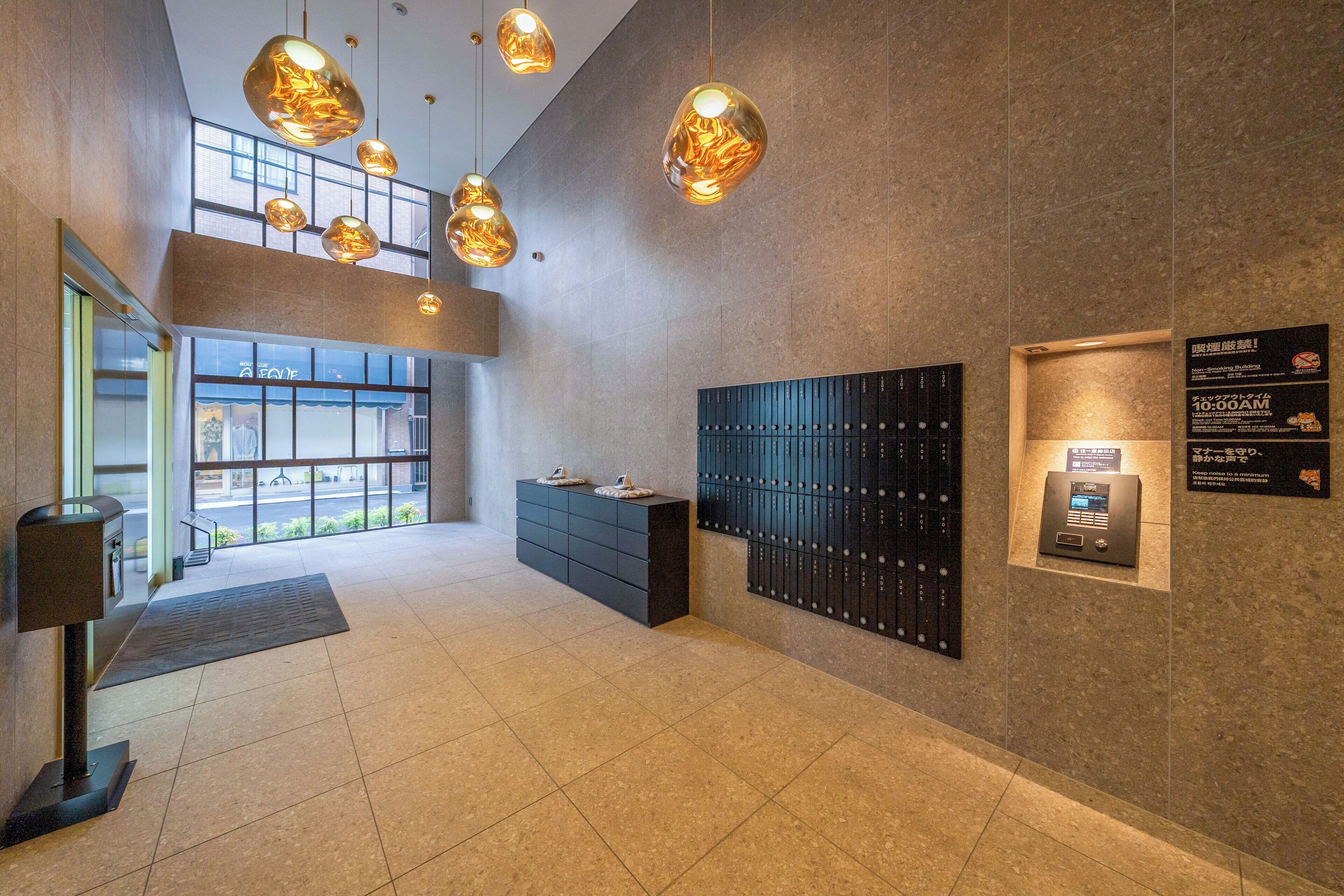 A modern lobby with a high ceiling, pendant lighting, a reception desk, a wall-mounted safe, and a view of the street through large windows.