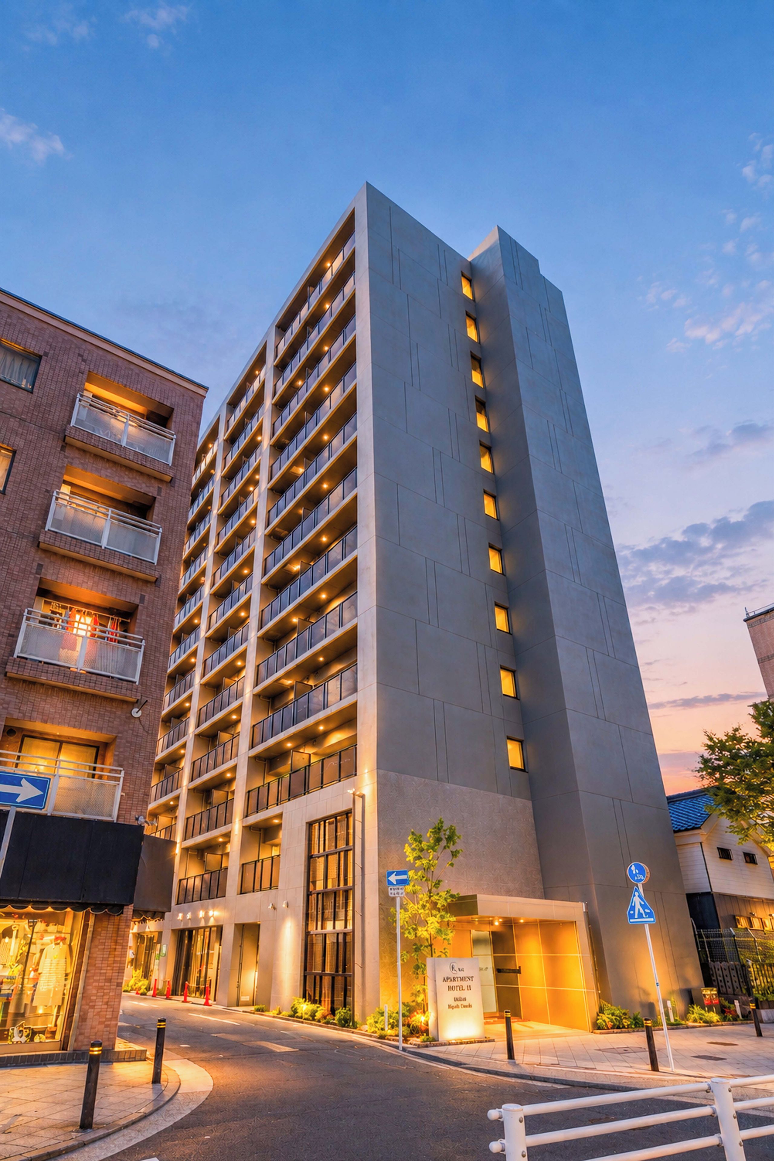 A multi-story hotel with a modern design, featuring balconies on each floor, a ground-level entrance, and a clear sky in the background.