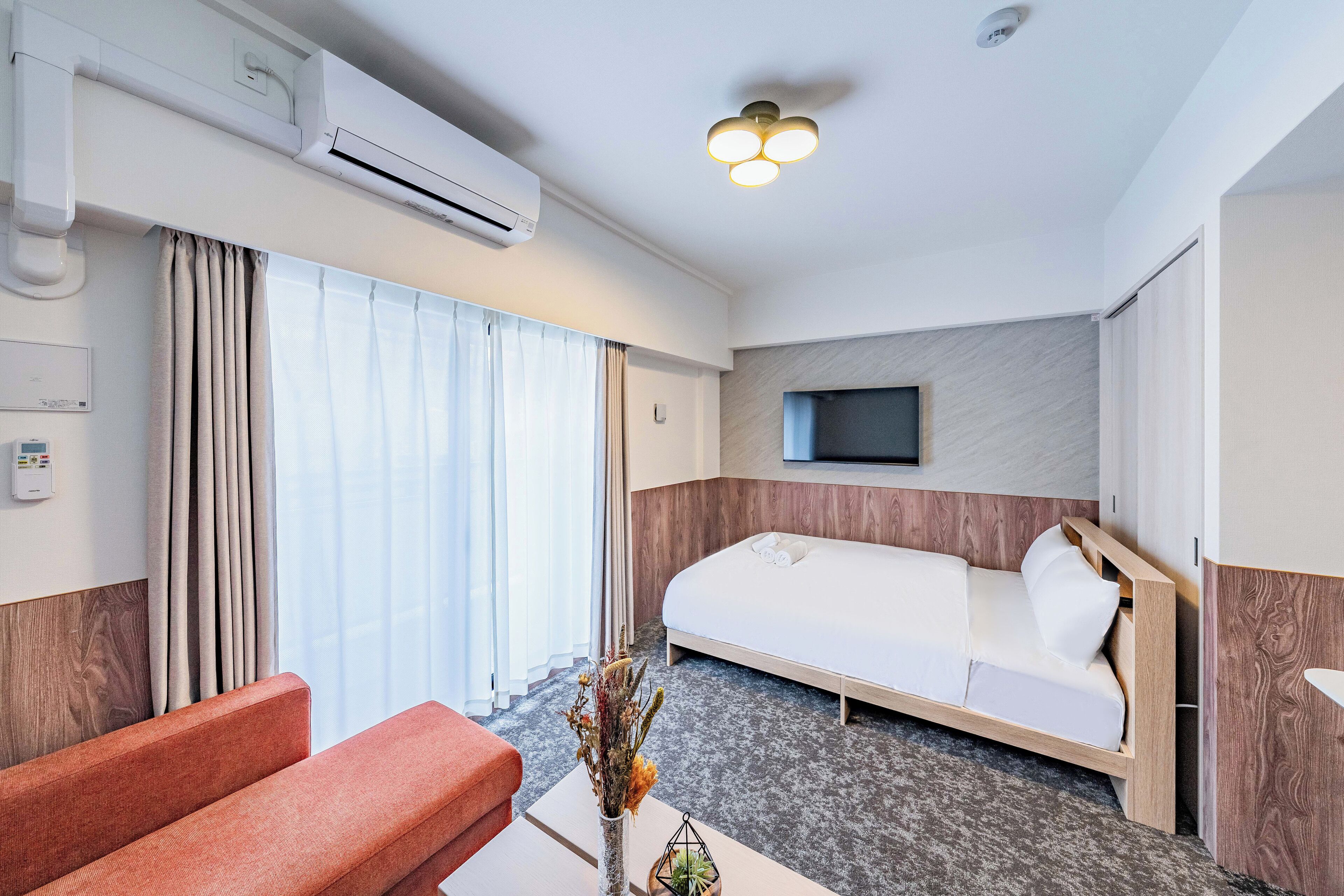 A hotel room with a bed, a sofa, a TV mounted on the wall, and an air conditioning unit.
