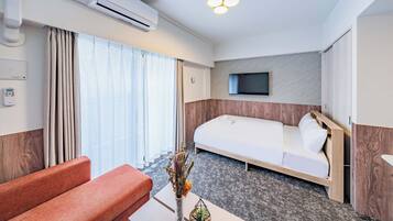 A hotel room with a bed, a sofa, a TV mounted on the wall, and an air conditioning unit.
