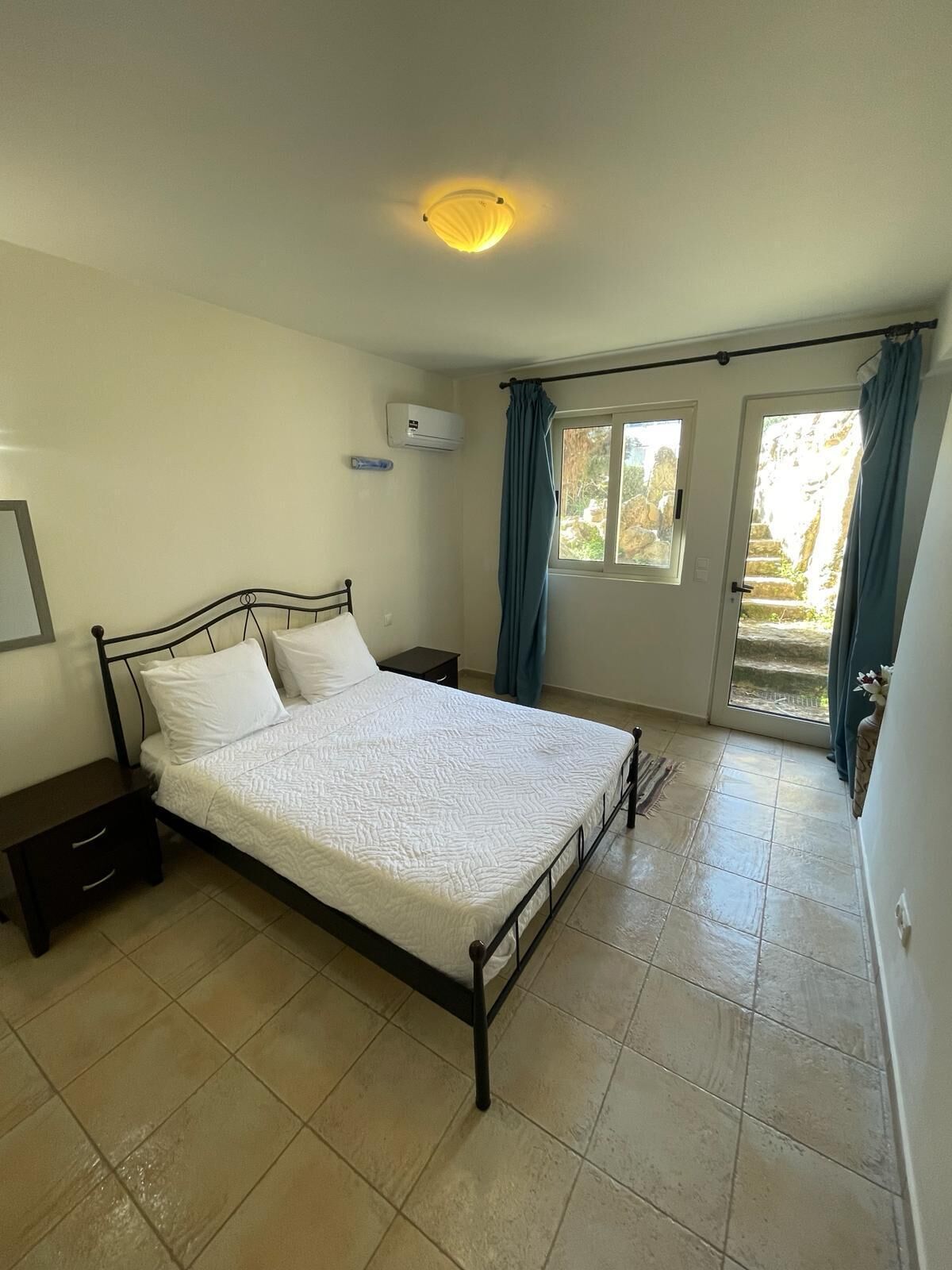 A bedroom with a bed, a nightstand, a window with curtains, and an air conditioning unit.