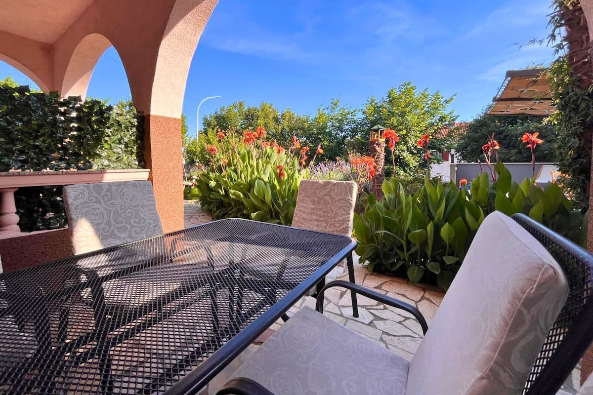 An outdoor patio with a metal table and chairs, surrounded by lush greenery and flowering plants.