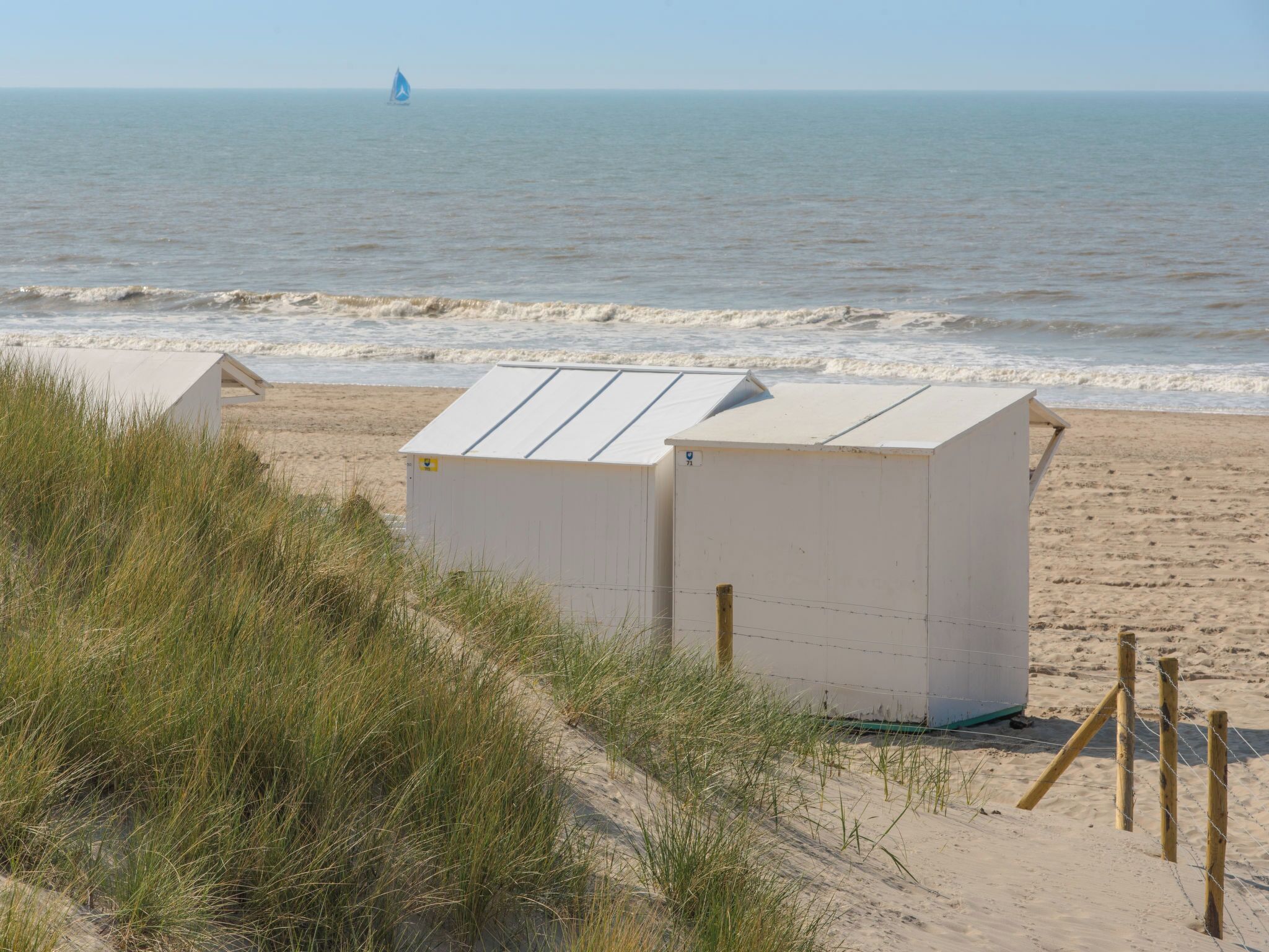 A beach with a dune system, a small white building, and a sailboat in the distance.