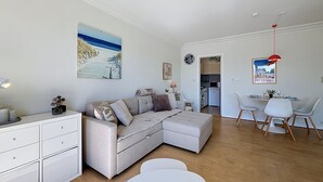 Una sala moderna con un sofá seccional, un comedor con sillas blancas, una cocina al fondo y un cuadro con temática playera en la pared.