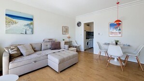 Una sala de estar moderna con un sofá modular, un comedor con mesa y sillas, una cocina al fondo y un cuadro con motivos playeros en la pared.