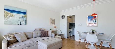 Una sala de estar moderna con un sofá modular, un comedor con mesa y sillas, una cocina al fondo y un cuadro con motivos playeros en la pared.
