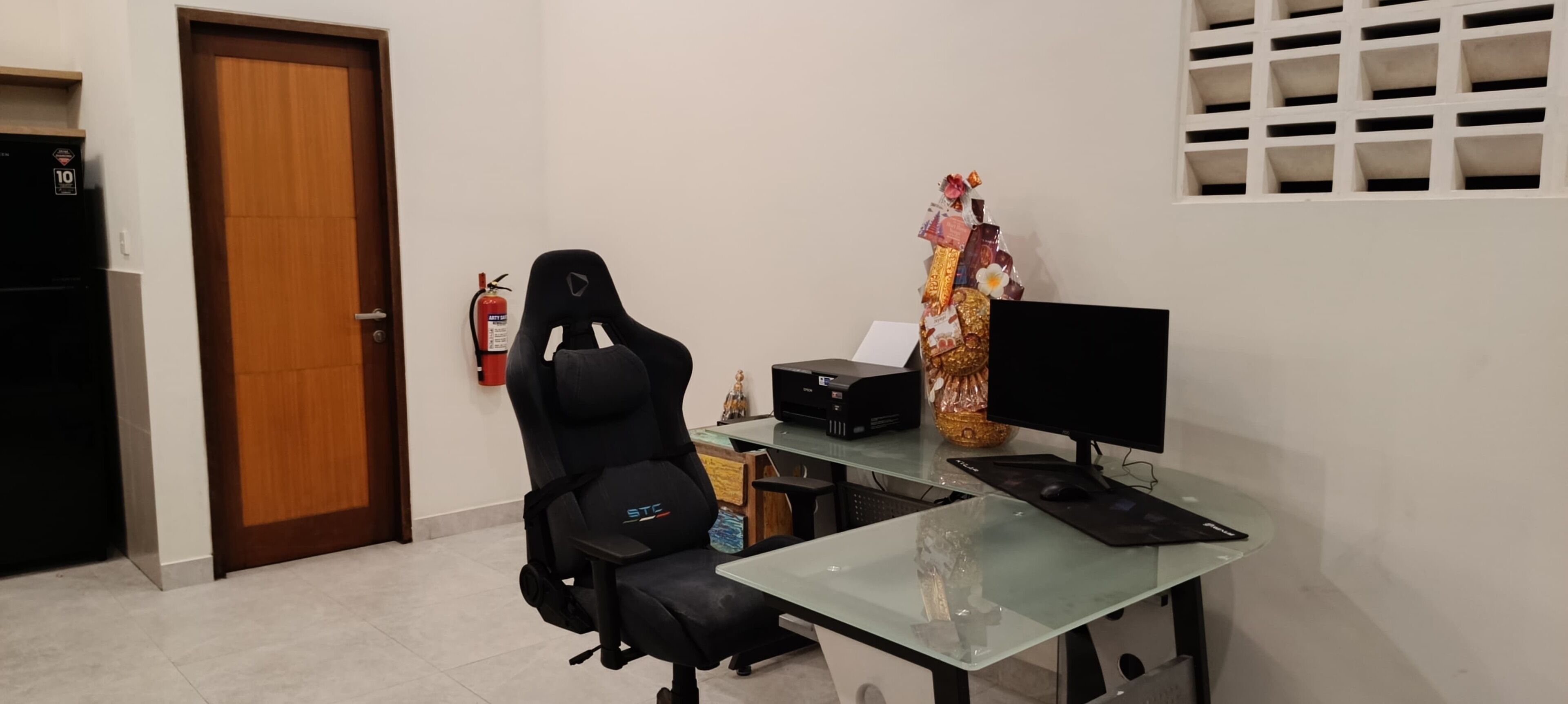 A room with a wooden door, a black gaming chair, a glass desk with a computer monitor, keyboard, and mouse. There is a fire extinguisher on the wall and a decorative item on the desk.