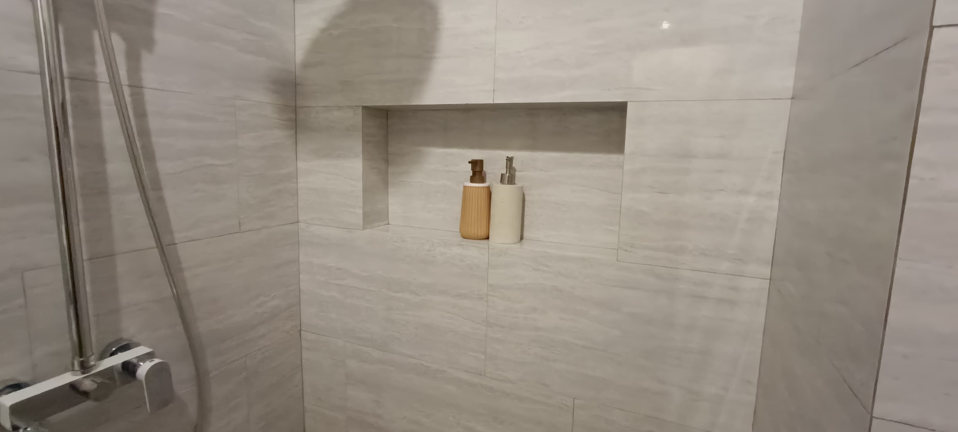 A modern bathroom with a glass shower enclosure, a built-in shelf with toiletries, and a marble-tiled wall.