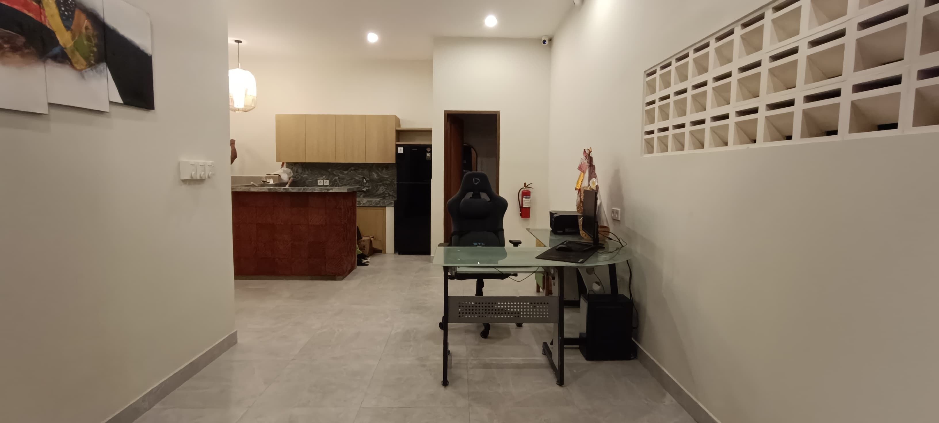 A narrow hallway with a kitchen area on the left, a desk with a computer and a chair on the right, and a white wall with a grid pattern on the far right.