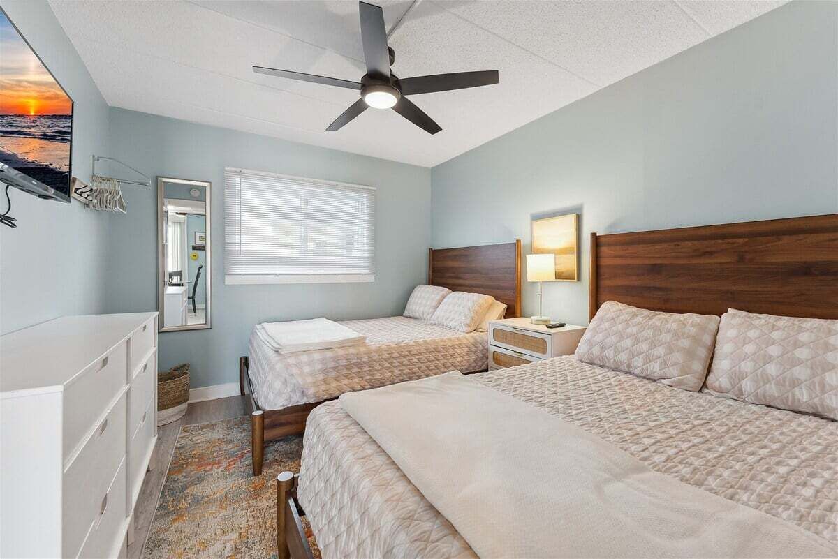 A bedroom with two beds, a ceiling fan, a window with blinds, a bedside table with a lamp, and a dresser.