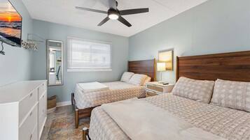 A bedroom with two beds, a ceiling fan, a window with blinds, a bedside table with a lamp, and a dresser.