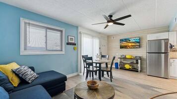 A living room with a sofa, a round table, a ceiling fan, and a kitchen area with a refrigerator.