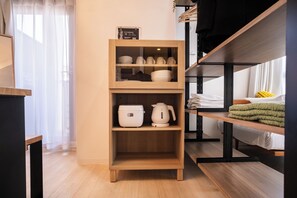Ein kleines Zimmer mit einer Holzeinrichtungsregal, das Geschirr enthält, einem Bett mit grüner Decke und einem Fenster mit transparenten Vorhängen.