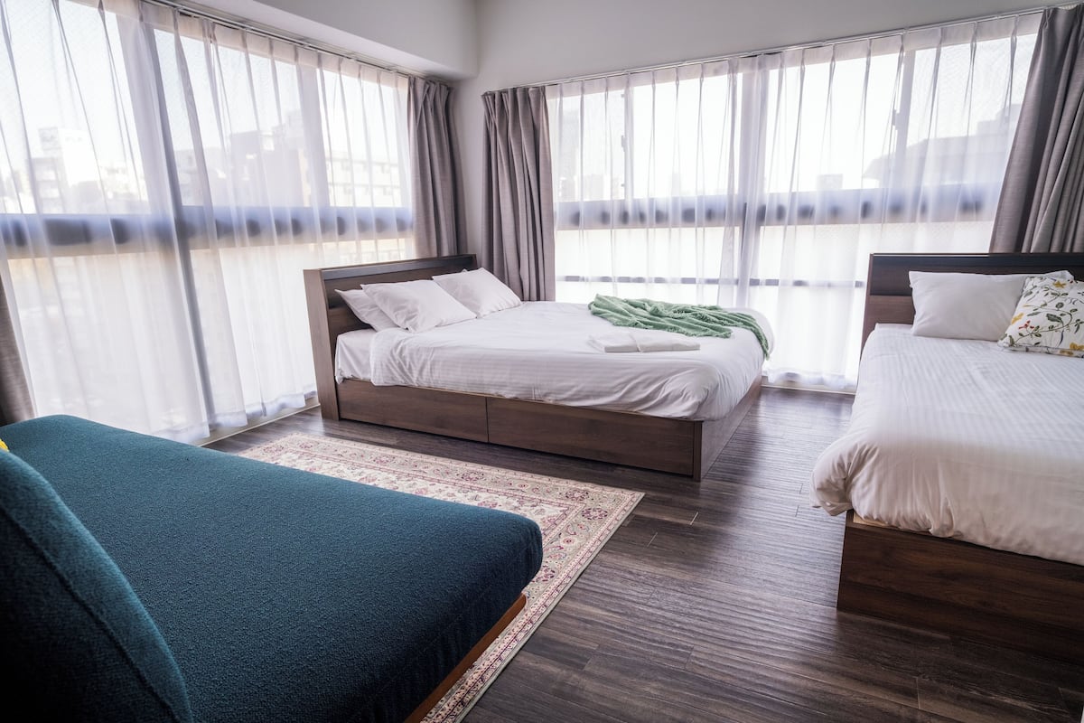 Ein Hotelzimmer mit zwei Betten, einem großen Fenster mit Vorhängen, einem Holzboden und einem grünen Sofa.