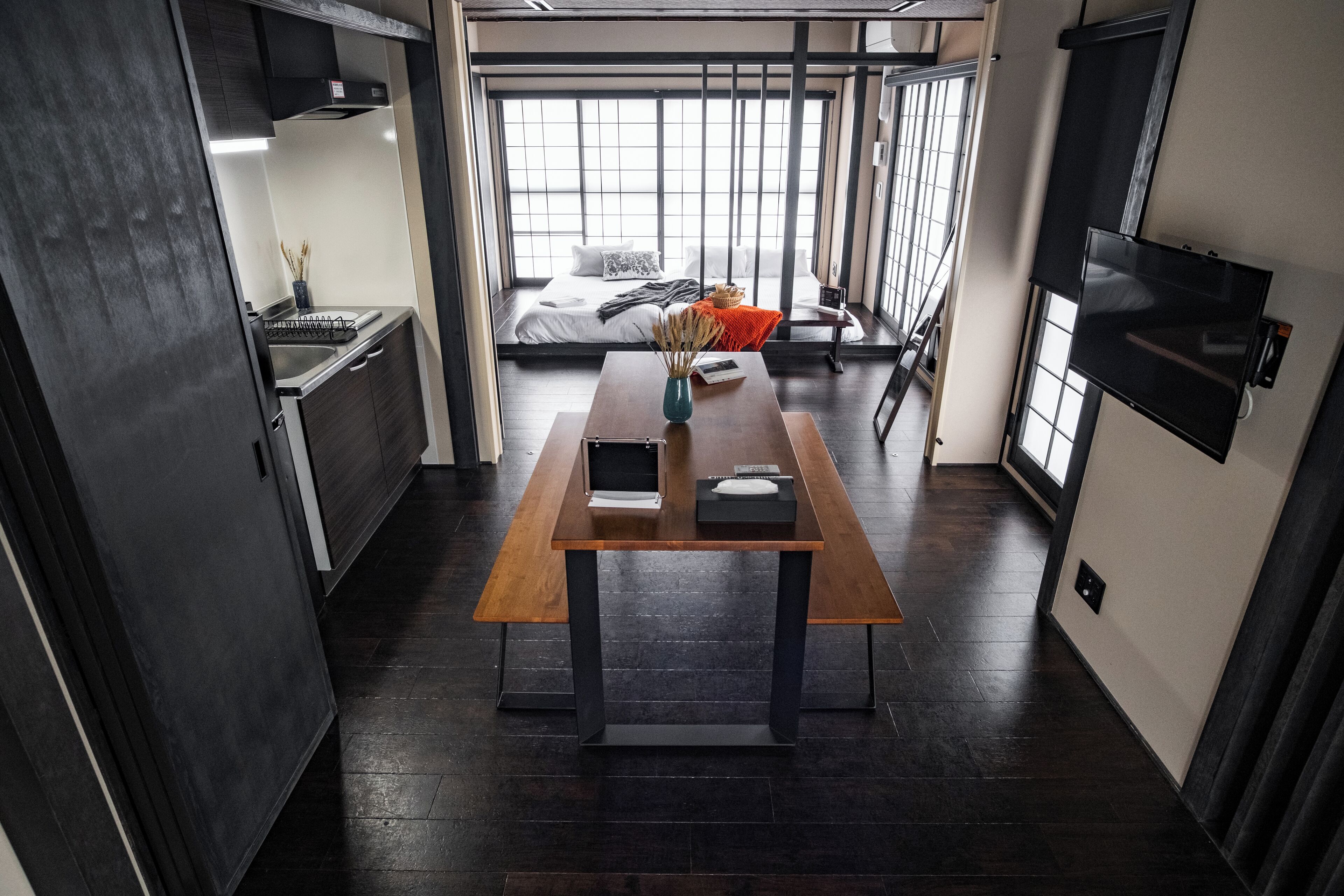 A modern hotel room with a wooden dining table, a bed with a view, a TV, and a kitchen area.
