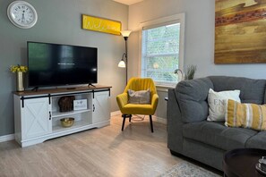 A living room with a flat-screen TV, a yellow armchair, a grey sofa, and a window with blinds. - Cute and Cozy 2 bedroom bungalow in Seminole Heights (Tampa)