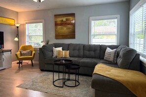 A living room with a gray sofa, yellow armchair, and a coffee table. There are two windows with blinds, a painting on the wall, and a lamp.