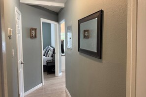 A hallway with light gray walls, white trim, and a wooden floor. There are two framed pictures on the walls, a door on the left, and a bed with pillows in the background.