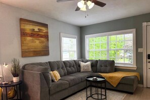 Ein Wohnzimmer mit grauem Sofa, Deckenventilator und großem Fenster mit Blick auf Grünflächen.