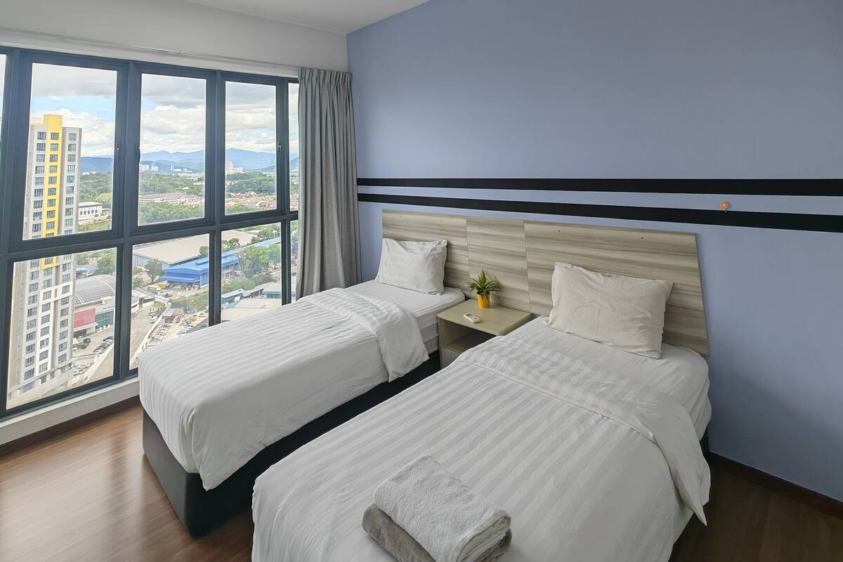 A hotel room with two beds, a large window with a city view, a wooden headboard, and a small bedside table with a plant.